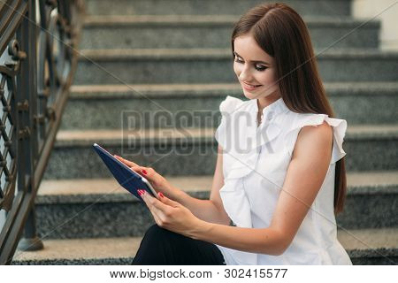 Young And Attractive Female Sittind On The Stairs And Use Tablet. Woman Play Online Games On Tablet