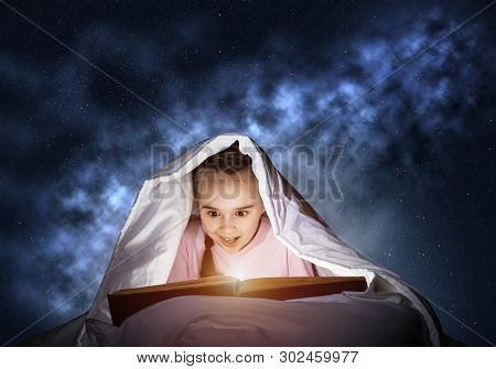 Engrossed Little Girl Reading Book In Bed Before Going To Sleep. Pretty Kid With Flashlight Hiding U