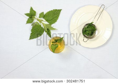 Healing Tea With Fresh Nettles, Flat View. Glass Of Nettle Tea, Tea Strainer On The Plate,  Light Ta
