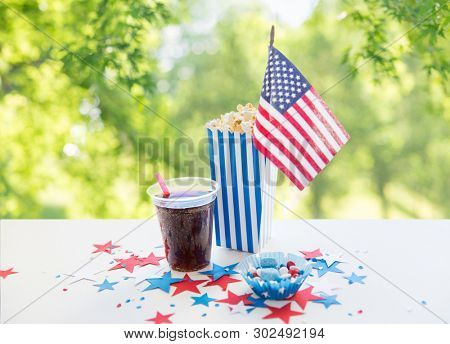 celebration and holidays concept - close up of american flag, cola cup, popcorn and candies with stars confetti decoration at 4th july party on independence day over green natural background