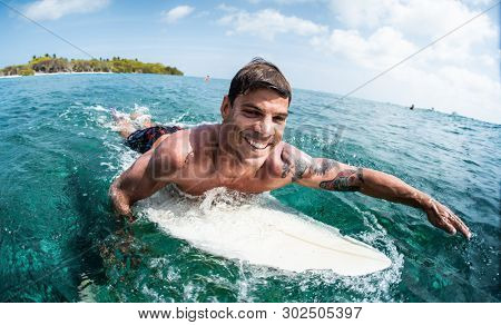 HIMMAFUSHI / MALDIVES - MARCH 10, 2019: Hispanic happy surfer paddles in the tropical ocean.