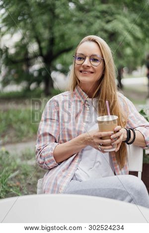 Blonde In A Coffee Shop At A Table On The Street. Paper Cup Of Coffee In Hand. Coffe To Go