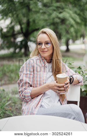 Blonde In A Coffee Shop At A Table On The Street. Paper Cup Of Coffee In Hand. Coffe To Go