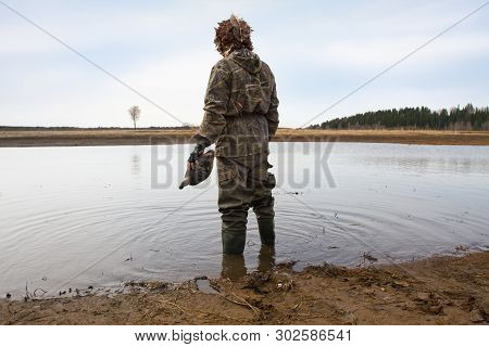 Duck Hunter With A Duck Decoy In His Hand Stands On The Shallow Water