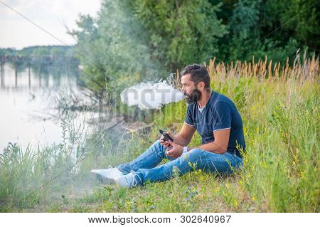 Confident Smoker Resting On The Nature And Smoking Electronic Smoke Device. Vaping Concept.