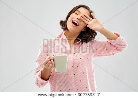 people and bedtime concept - happy sleepy young woman in pajama with mug of coffee yawning over grey background