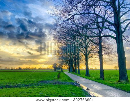 Dramatic And Colorful Sunrise Or Sunset Sky Over A Grassy Green Farmfield Lined By Trees
