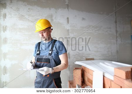Male Builder In Yellow Hard Hat Holds Clipboard On Background Of An Apartment Under Repair Portrait.