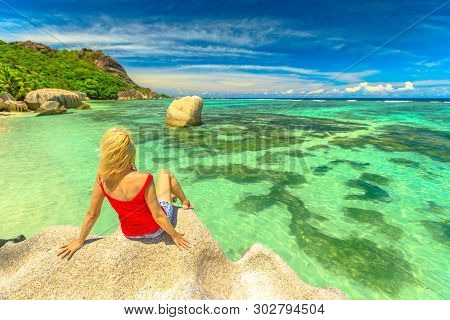 Tourist Woman Sitting On A Granite Boulder At Anse Source Dargent. Blonde Girl Looks Turquoise Sea O