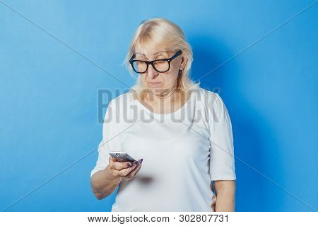 Old Woman Is Holding A Tv Remote On A Blue Background. Concept Television For The Elderly, The Study