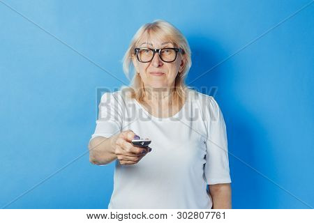 Old Woman Is Holding A Tv Remote On A Blue Background. Concept Television For The Elderly, The Study