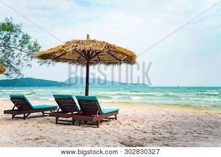 Two Chaise Longues Under A Straw Umbrella On A Beach Near The Sea. Tropical Background. Coast Of Isl