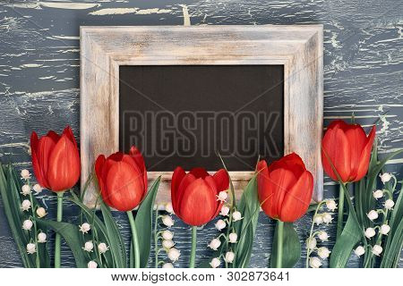Blackboard And Bunch Of Red Tulips And Lily Of The Valley Flowers On Rustic Background. Space For Yo