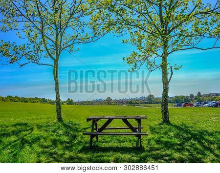 Single Empty Wooden Picnic Table Set Between Two Trees On Grass Looking Towards The View Of London.