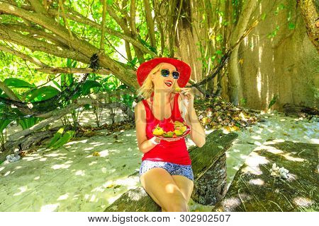 Tourist Woman In Red Sunhat Eats Fresh Local Fruit Plate At Anse Source Dargent, La Digue Under Palm