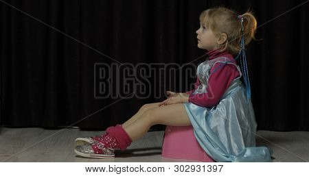 Little Girl Sitting On The Potty And Watching Tv. Blonde Cute Child In Blue Dress. Inside