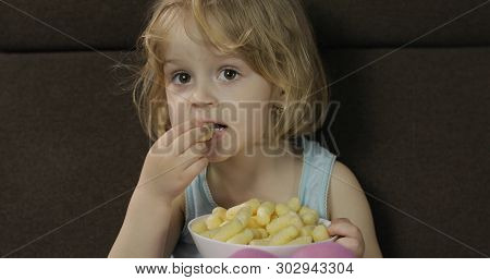 Beautiful Little Blonde Girl Sitting On A Sofa At Home And Eating Corn Puffs. Cute Child Taste Puffc