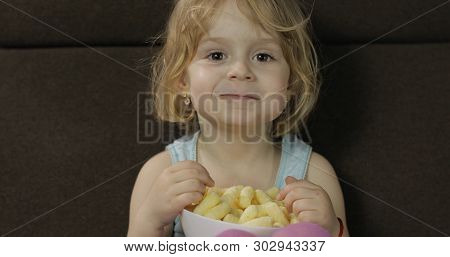 Beautiful Little Blonde Girl Sitting On A Sofa At Home And Eating Corn Puffs. Cute Child Taste Puffc