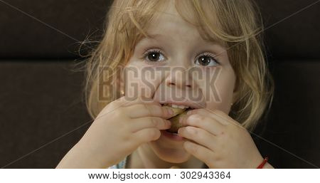 Beautiful Little Blonde Girl Sitting On A Sofa At Home And Eating Corn Puffs. Cute Child Taste Puffc
