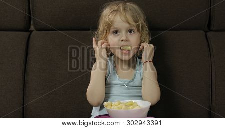 Beautiful Little Blonde Girl Sitting On A Sofa At Home And Eating Corn Puffs. Cute Child Taste Puffc