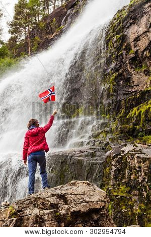 Tourist Woman With Norwegian Flag At Svandalsfossen In Norway, Powerful Waterfall In Norwegian Mount