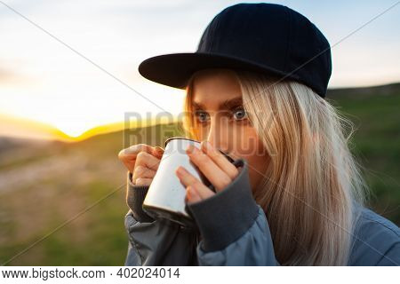 Close-up Portrait Of Young Blonde Girl With Black Cap Drinking Hot Tea From Steel Mug In Background 