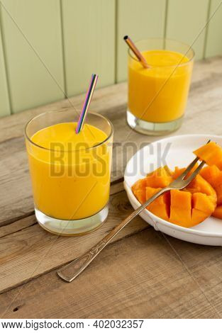 Fresh Mango Lassi In Two Glasses With Metal Straws And Sliced Mango Fruit With Fruit Horn Fork On Wo