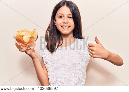 Beautiful child girl holding potato chip smiling happy and positive, thumb up doing excellent and approval sign 