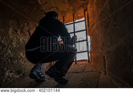 Man  Traped In A Dungeon Holding The Bars Of A Small Window An Looking Outside