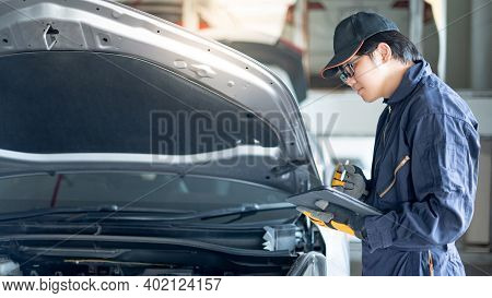 Asian Auto Mechanic Holding Digital Tablet Checking Car Engine Under Bonnet Hood In Auto Service Gar