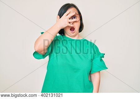 Brunette woman with down syndrome wearing casual clothes and glasses peeking in shock covering face and eyes with hand, looking through fingers afraid 