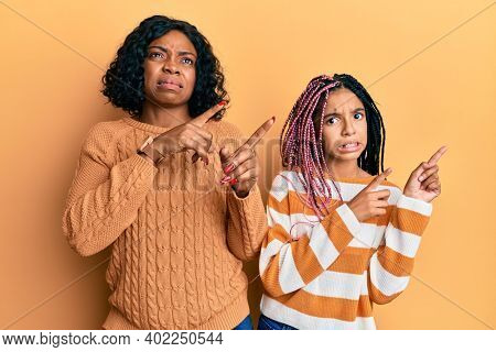 Beautiful african american mother and daughter wearing wool winter sweater pointing aside worried and nervous with both hands, concerned and surprised expression 