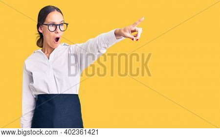 Beautiful brunette young woman wearing professional waitress apron pointing with finger surprised ahead, open mouth amazed expression, something on the front 