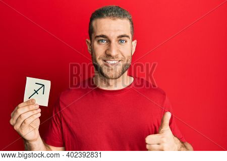 Young caucasian man holding paper with sagittarius zodiac sign smiling happy and positive, thumb up doing excellent and approval sign 