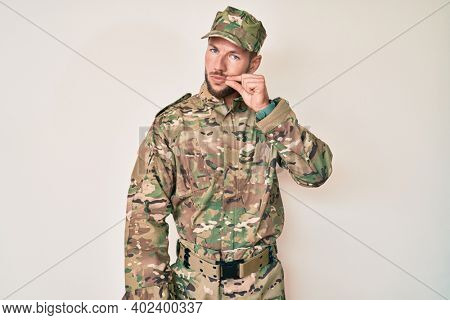 Young caucasian man wearing camouflage army uniform mouth and lips shut as zip with fingers. secret and silent, taboo talking 