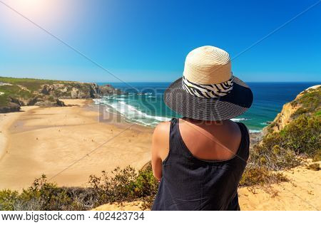 A Woman With A Hat On Her Head, On The Shore, Admires The View Of The Portuguese Beach, Odeceixe.