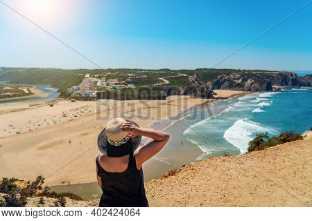 A Woman With A Hat On Her Head, On The Shore, Admires The View Of The Portuguese Beach, Odeceixe.