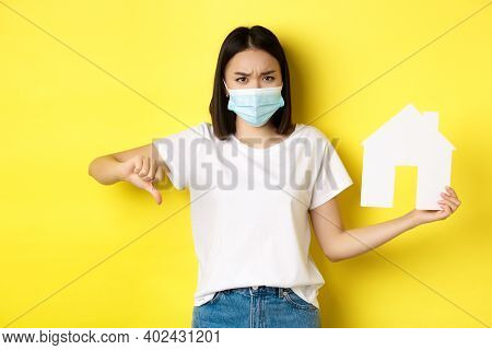 Covid-19 And Real Estate Concept. Disappointed Asian Woman In Medical Mask, Showing Thumb Down And P