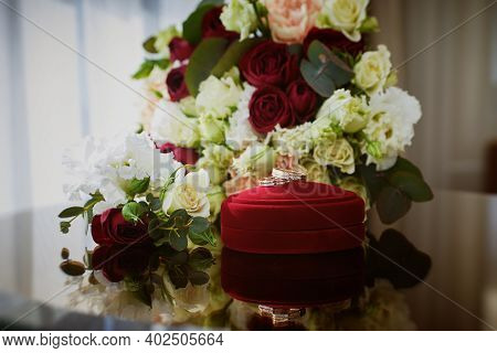 Close Up View Of Wedding Rings On A Red Velvet Box And Flowers On A Glass Table In The Hotel Room.