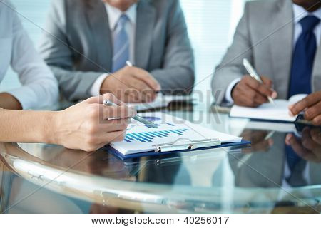 Detail lidské ruky s perem, ukázal na papír zároveň něco vysvětlovat kolegy