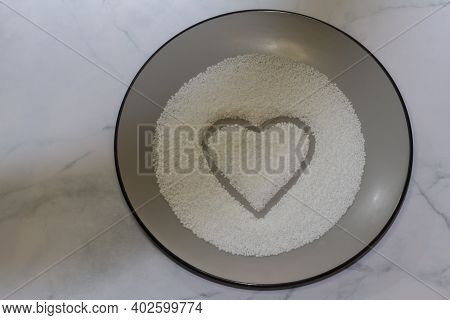 Pearl Sugar Is Poured Into A Gray Plate In The Shape Of A Heart. White Gray Marble Rustic Background