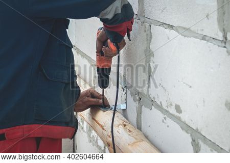 A Male Worker In Overalls Uses An Electric Screwdriver To Screw Screws Into A Wooden Beam. Construct