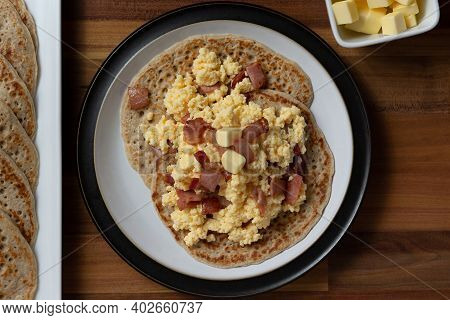 Scrambled Egg And Bacon Stacked On Top Of Derbyshire Oatcakes On A Black Rimmed Plater. A Stack Of O