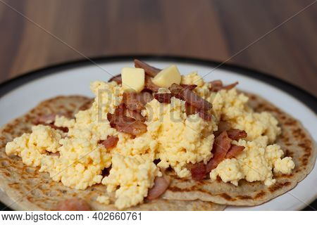 A Close Up View Of Derbyshire Oatcakes Served With Scrambled Eggs And Bacon On A Black Rimmed Plate