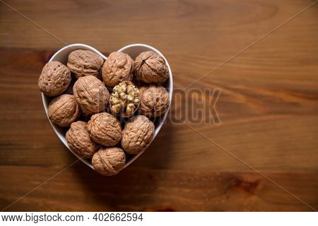 Whole Walnuts And One Cracked Broken Heart In A White Heart-shaped Bowl On A Wooden Background. Top 