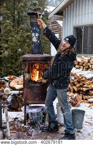 Young Man Playing With A Forge And Raising A Hammer, Laughing And Pretending To Be A Blacksmiths App