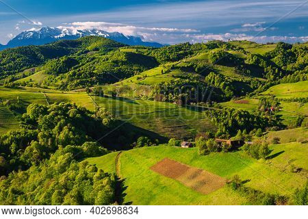 Picturesque Spring Countryside Scenery With Houses On The Hills And High Snowy Mountains In Backgrou
