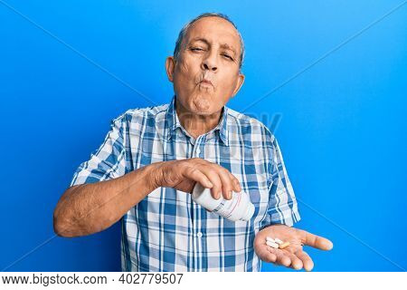Senior hispanic man holding pills making fish face with mouth and squinting eyes, crazy and comical. 