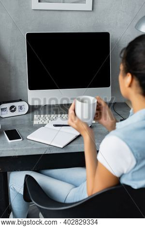 Young African American Freelancer Holding Cup Of Tea Near Computer Monitor, Smartphone And Notebook 