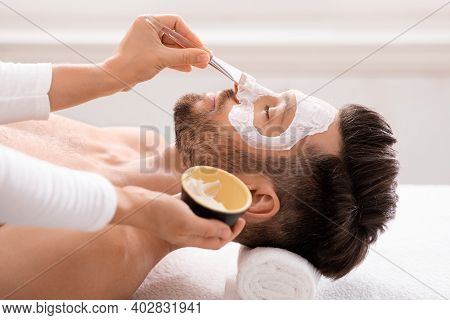 Side View Of Bearded Caucasian Man Getting Face Treatment At Modern Spa Salon. Female Attendant Appl